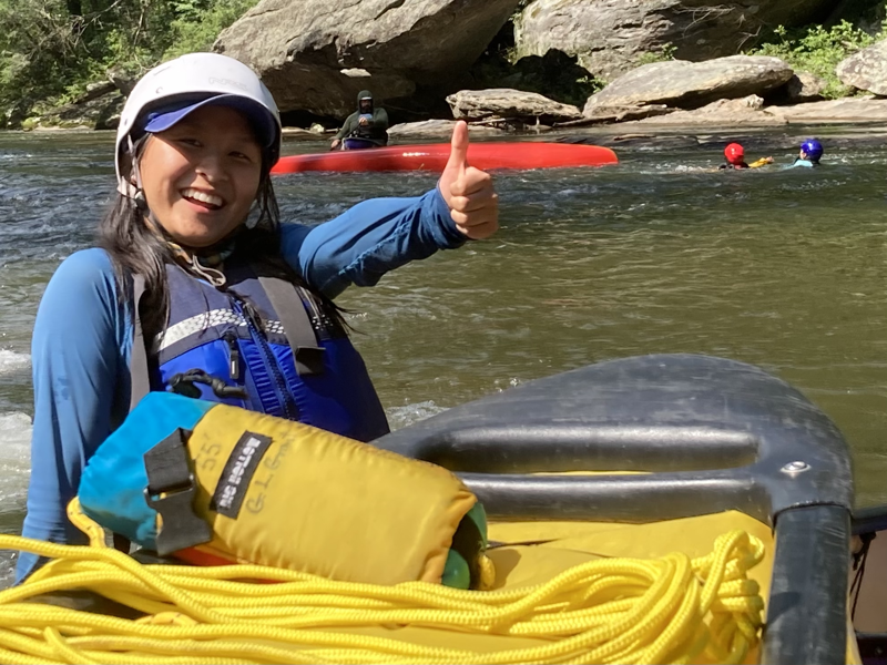 A smiling woman in a white helmet and blue life vest gives a thumbs up while sitting in a raft with yellow rope and a rescue throw bag. In the background, other people are kayaking and swimming in a river with large rocks on the banks. The scene suggests a whitewater rafting or kayaking adventure on a sunny day.